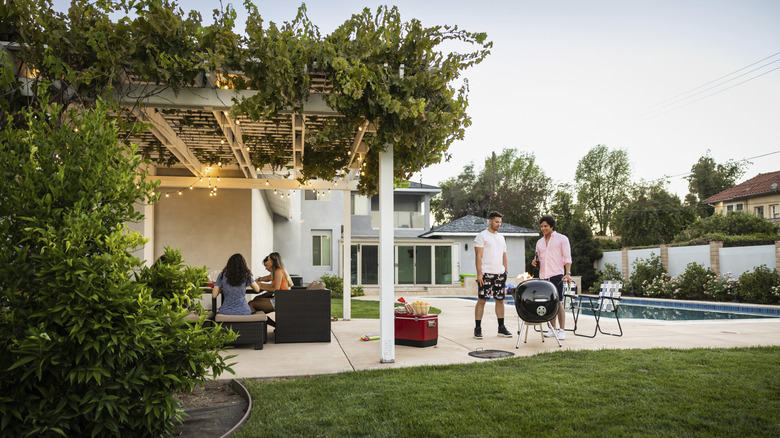 Guests sit under a vine-covered pergola in a backyard while two people use a BBQ beside a swimming pool.