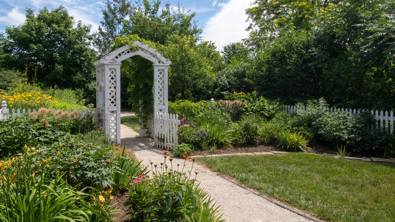 A white painted wood arbor over a gravel path between two gardens.