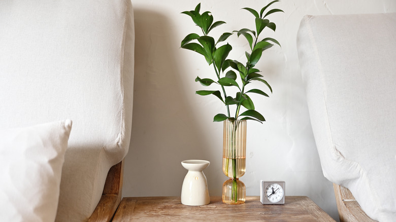 A curated end table between two sofa chairs displaying an oil burner, a tall vase with foliage, and a small clock