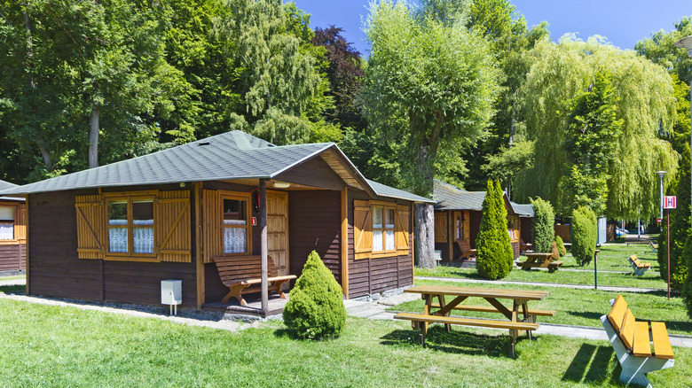 Rows of cabins at summer camp, picnic benches and shrubs surrounding them.