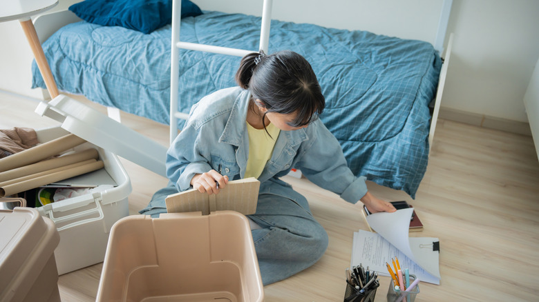 A woman sits on the floor in a college dorm room surrounded by school supplies.