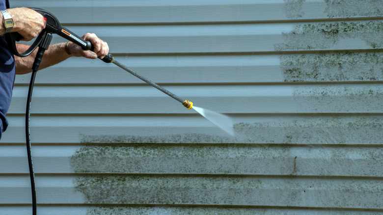 A person uses a pressure washer jet spray to clean mold off vinyl siding