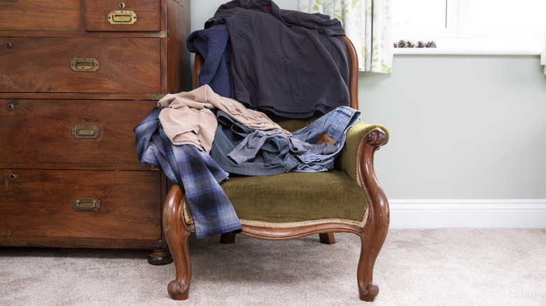 Pile of clothes on antique armchair with a wooden chest of drawer behind it.