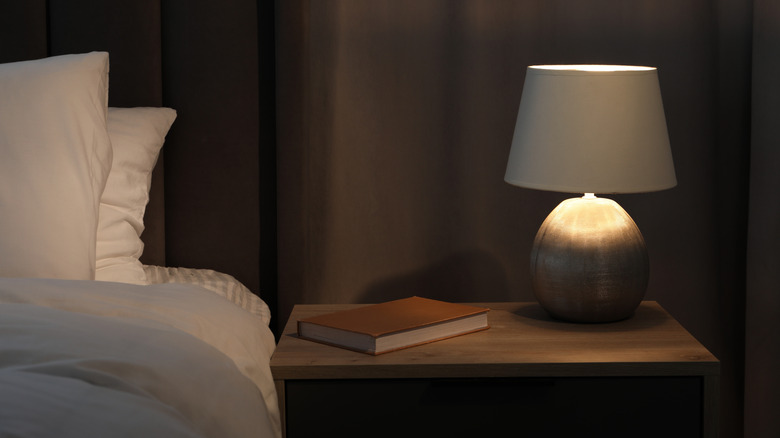 A light bedside lamp on a wooden nightstand with a book and the bed next to it.