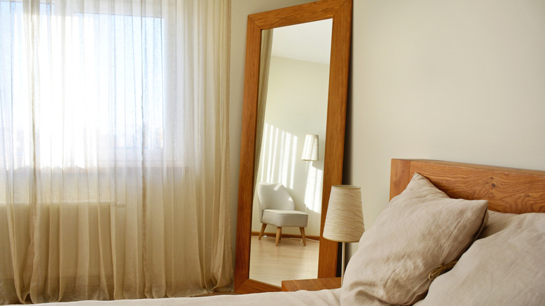 A large mirror in the corner of a bedroom with sheer curtains and a window next to it.