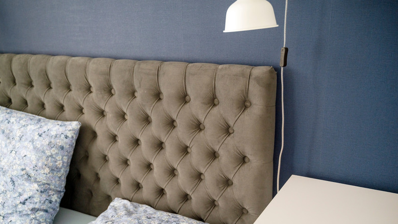 Close up of a quilted fabric headborad in brown with a blue wall and reading lamp behind it.