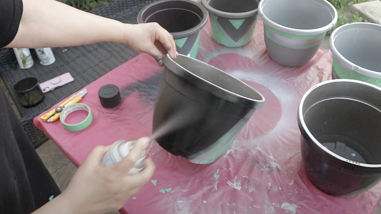 A person spray paints a plastic flower pot black outside on a table.