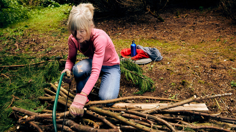 A mature light-skinned woman cuts branches with a hand saw.