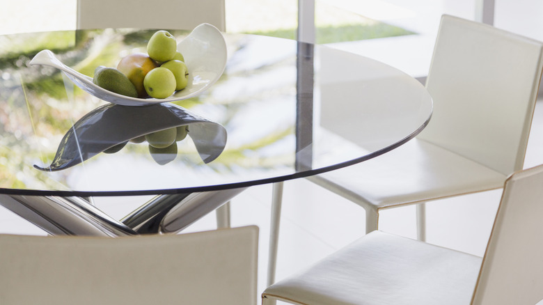 Close-up of a clean glass tabletop with a bowl of apples and white chairs around
