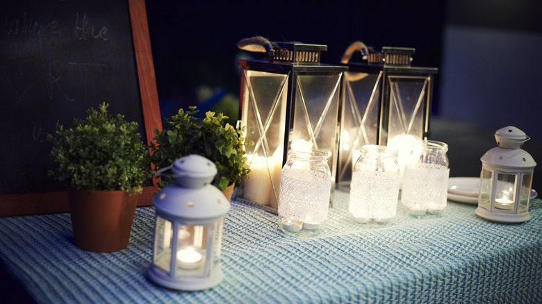 A collection of lighted outdoor table lamps are set up next to potted plants.