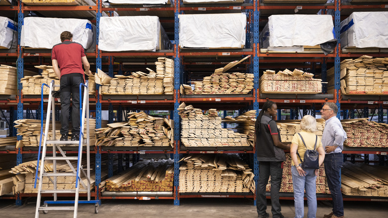 A couple standing with a worker in front of shelves full of lumber in a store.