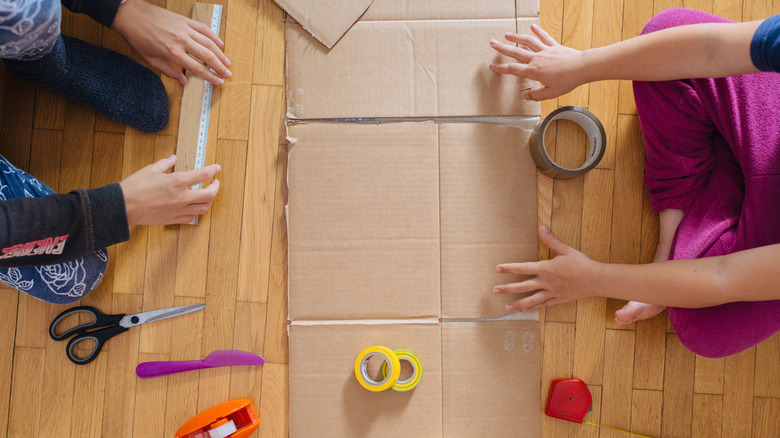 Two people working on a craft project on the floor using cardboard, tape, ruler, and scissors