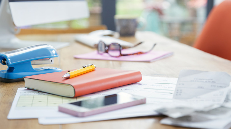 cluttered desk with phone, papers, log book, stapler, and glasses