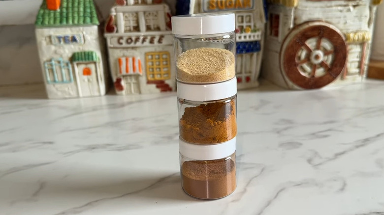Three small clear plastic jars storing spices stacked on top of each other on a granite countertop, a set of decorative jars that look like houses behind it
