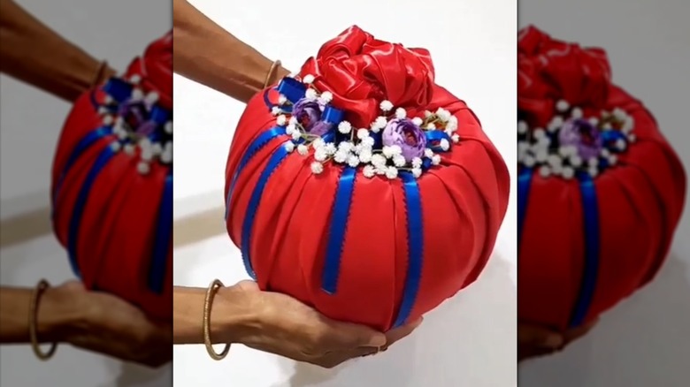 Black woman's hands holding a fish bowl made into a gift bag wrapped in red cloth, then decorated with sprigs of little white flowers and blue ribbon