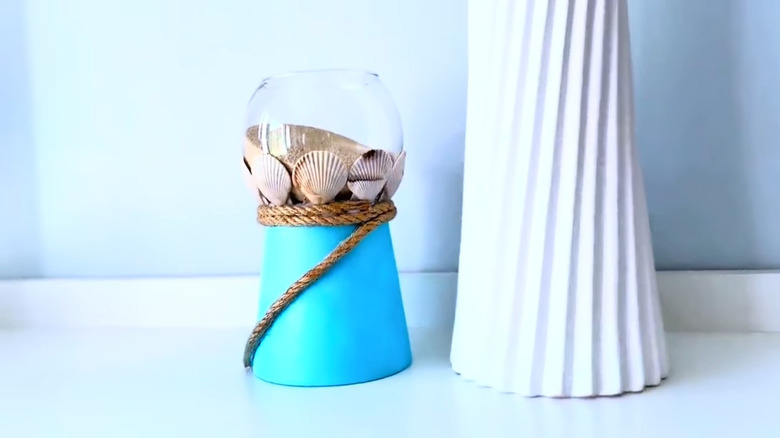 Light blue raised fish bowl with sea shell accents and rope on a pedestal next to a ribbed texture white vase