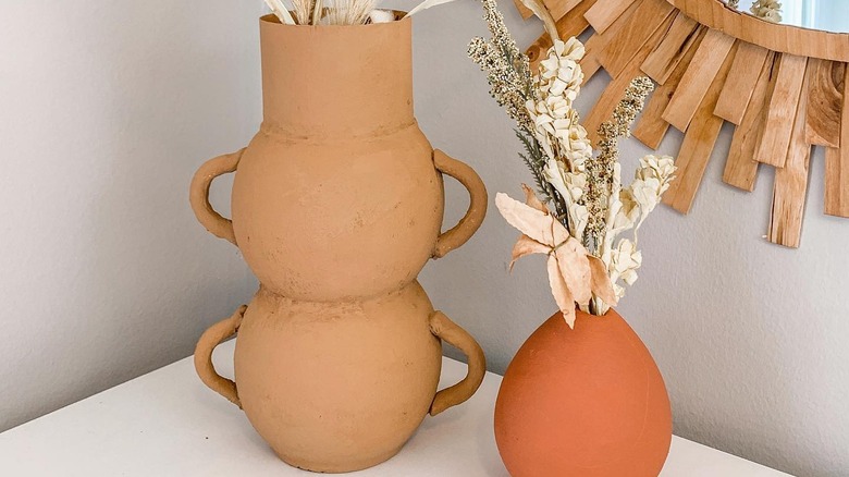 Tall textured vase in brown-orange color with handles sits on a counter next to an orange tear-shaped vase beside a mirror bordered with wood slats