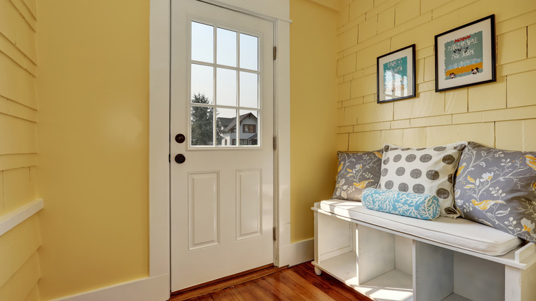 A front entry with yellow walls, a white door, prints on the wall, and a white bench with scatter cusions