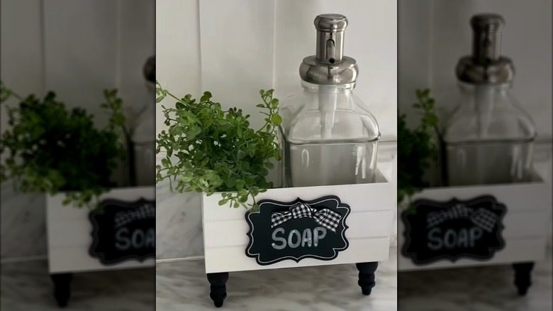 A bathroom soap pump holder and faux plant stored in a small, painted crate