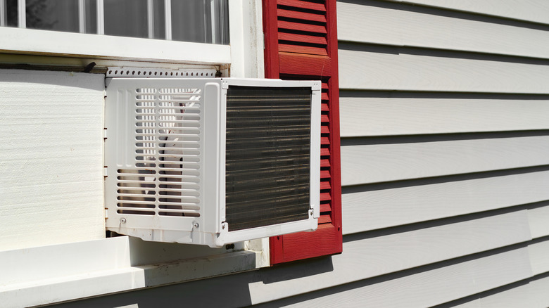 Window air conditioner sticking out of a window on a gray house with red shutters
