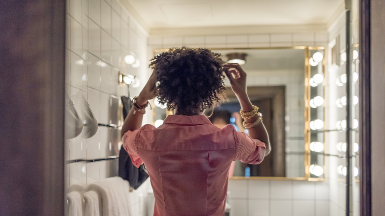 Woman fixing her hair in the mirror, bright vanity lights shine, white tile walls
