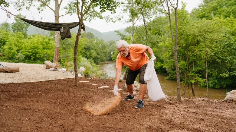 A man sows grass seeds over freshly prepared soil in his backyard near a river.