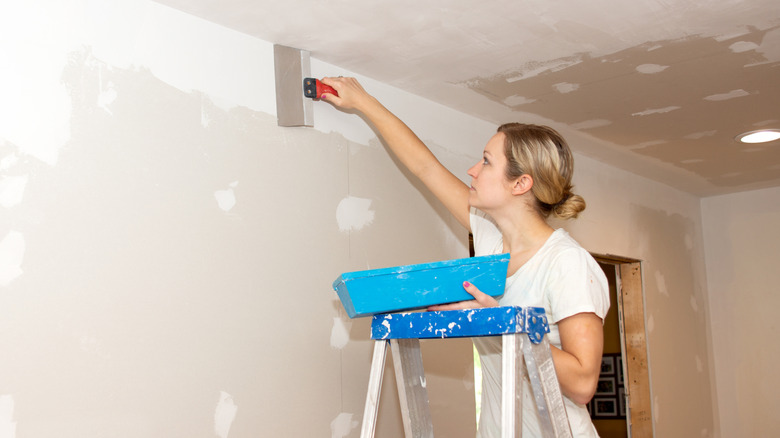 A woman stands on a ladder while smoothing out joint compound on newly hung drywall