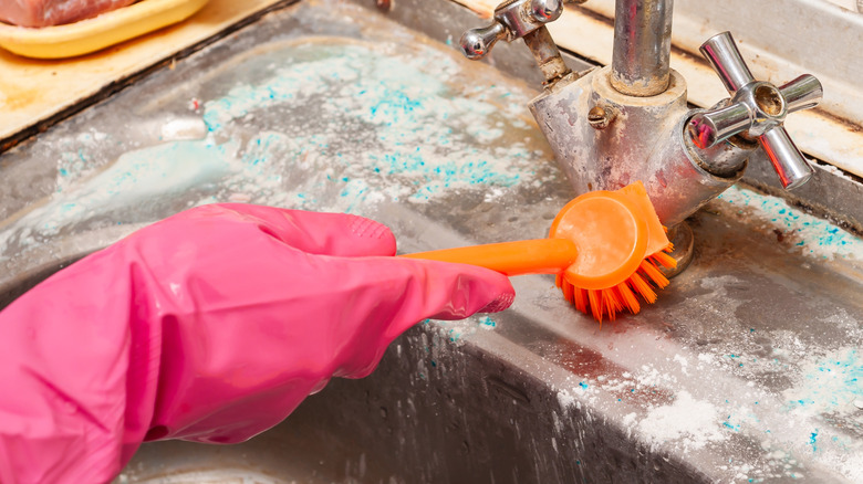 A pink rubber gloved hand holding an orange scrub brush scrubs soap scum off a dirty looking metal sink fixture