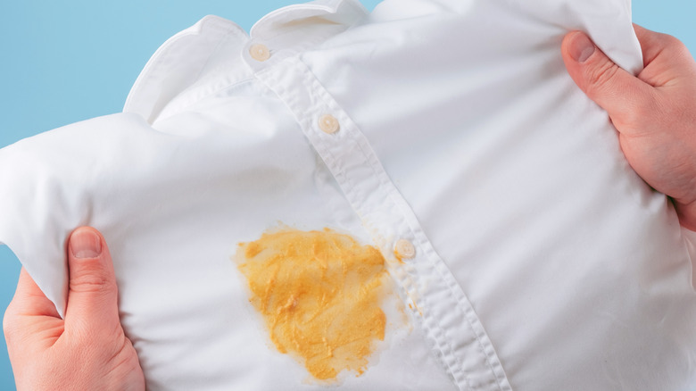 Hands holding up a white button down shirt with a large yellow mustard stain on it