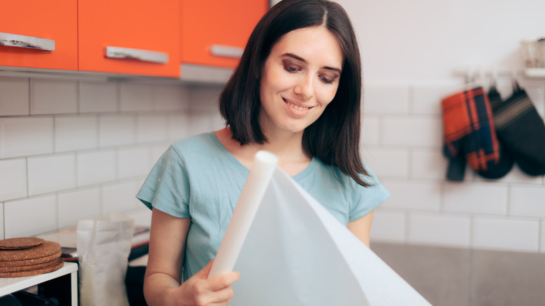 A smiling brown-haired woman unrolls a piece of wax paper in the kitchen.