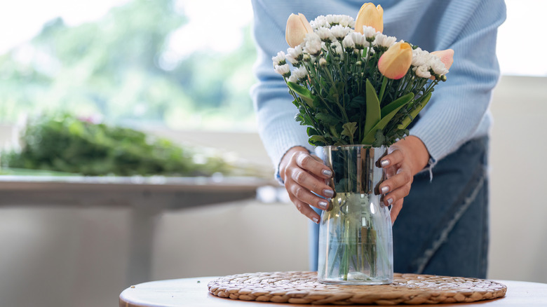 A person picking up a plain glass vase with flowers from a table