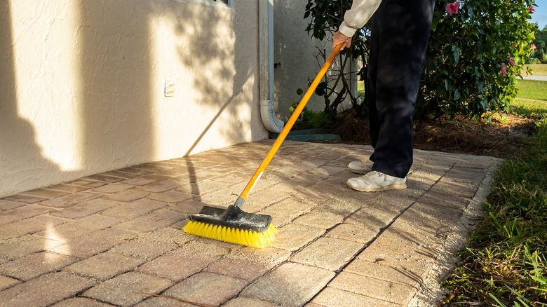 A person using a yellow push broom on outdoor pavers alongside a house