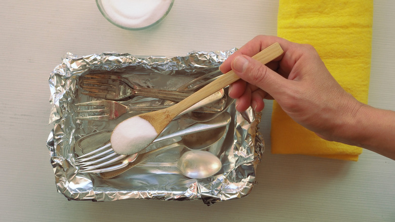 person putting spoonful of baking soda in aluminum-foil-lined and water-filled container holding silverware