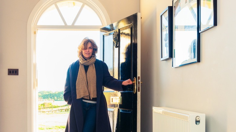 A short-haired, smiling woman enters a home through an arched doorway.