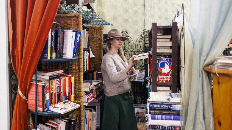 A woman wearing a hat browses the book selection in a cramped corner of a thrift store.