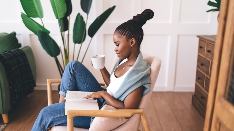 A woman reading a book and drinking coffee in side chair next to large living room plant