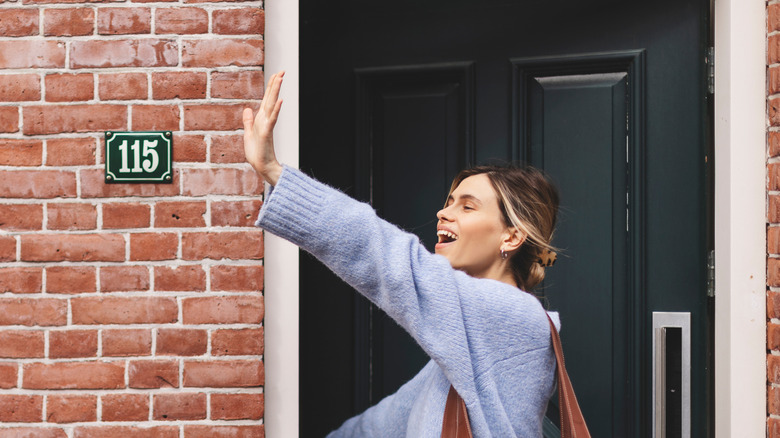 friendly, happy woman waving to someone, standing in a doorway next to a small house number