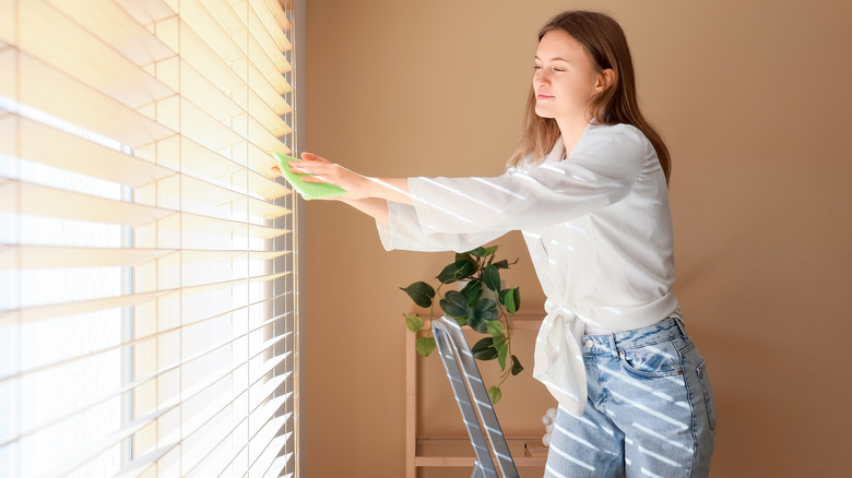 Woman on ladder dusting discolored window blinds