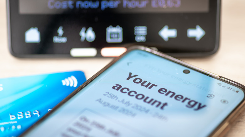 A phone showing a digital energy bill and a credit card sit in front of a home thermostat
