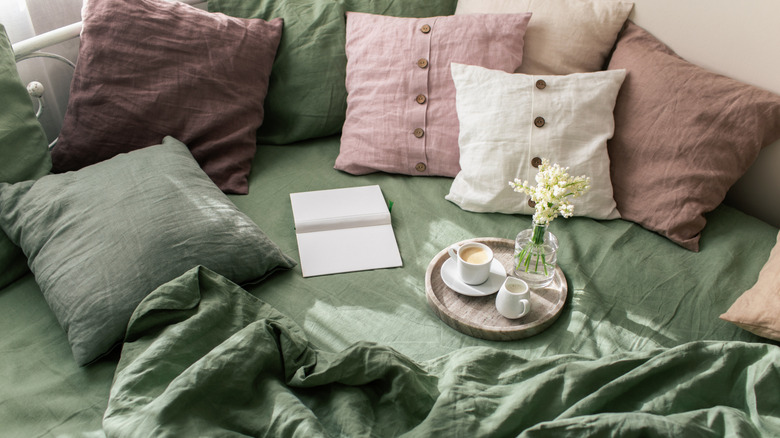 green linen sheets on bed with lots of pillows, a journal, and a tray with coffee and a vase of flowers