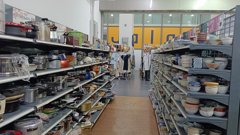 A kitchenware aisle in a thrift store with pots, pans, dishes, and more on shelves