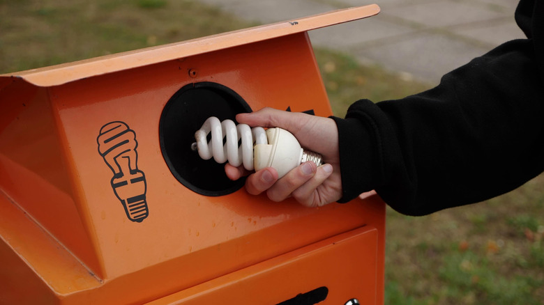 A hand placing a CFL light bulb in a recycling bin