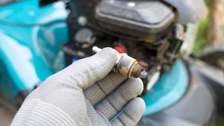 A gloved hand holds a corroded lawn mower spark plug above a blue lawn mower.