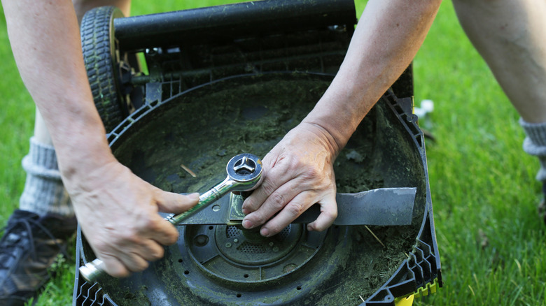 A person stands over a lawn mower as he uses a tool to fix it.