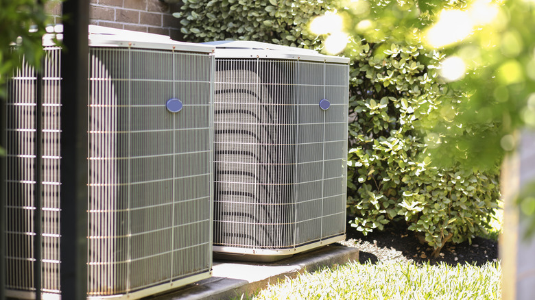 Two AC units outside by a brick wall with greenery on all sides.