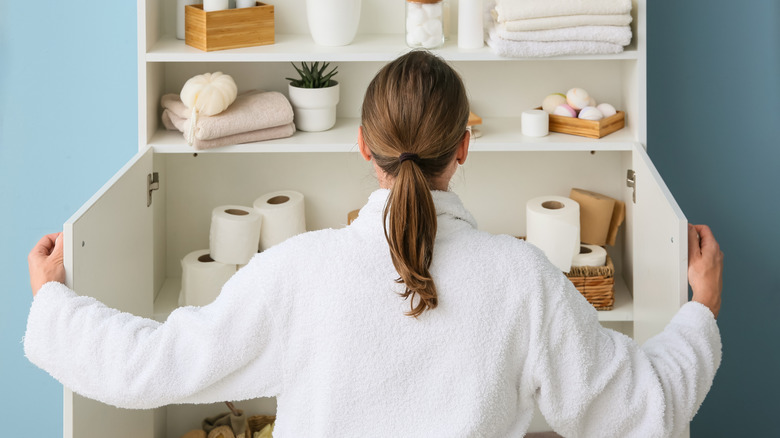 A woman looking at an open white bathroom cabinet with towels and bathroom essentials