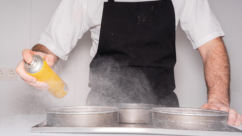 A cook using cooking spray on metal cake pans