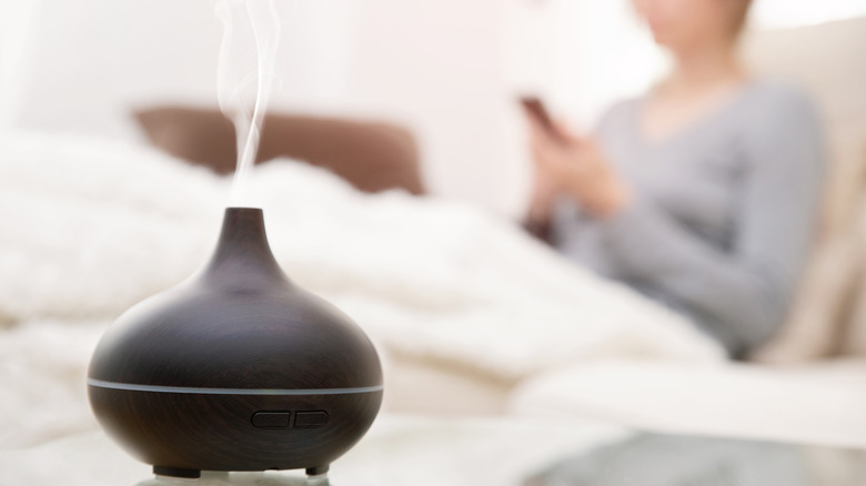 A black diffuser with mist coming out with the blurred image of a person sitting up in bed in the background