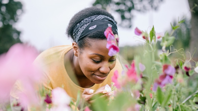 A Black woman wearing a black bandana and yellow shirt bends over to smell flowers, with pink and purple flowers blurred in the foreground.