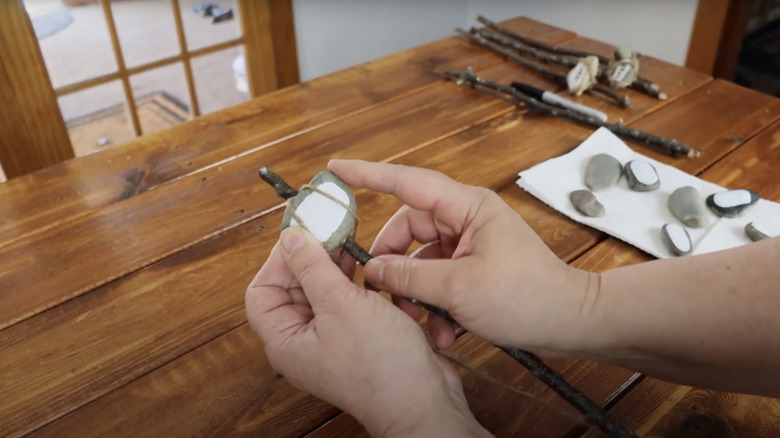 A crafter holds a painted pebble onto a twig as she wraps twine around it.